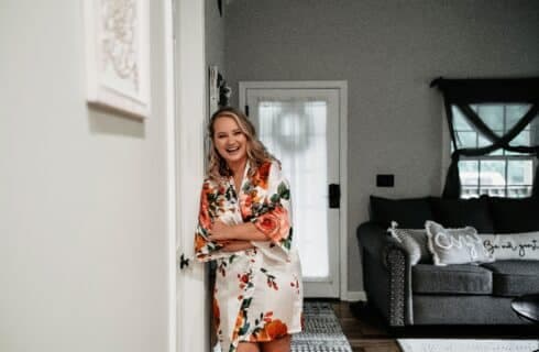 A smiling woman in a floral robe stands by an open door in a cozy living room.