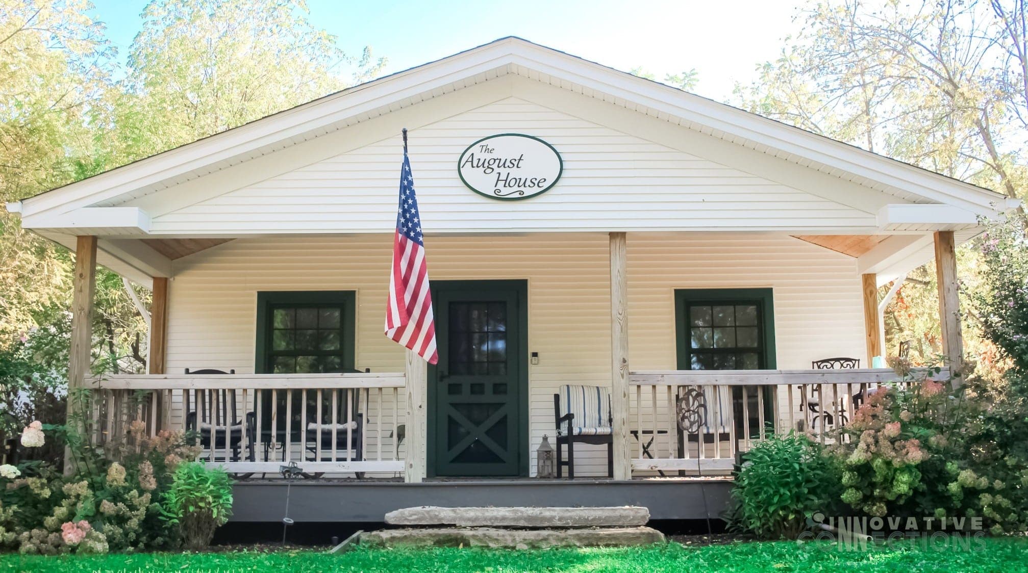 A white cottage with green trim and a front porch, featuring an American flag and a sign reading "The August House." A white cottage with green trim and a front porch, featuring an American flag and a sign reading "The August House."