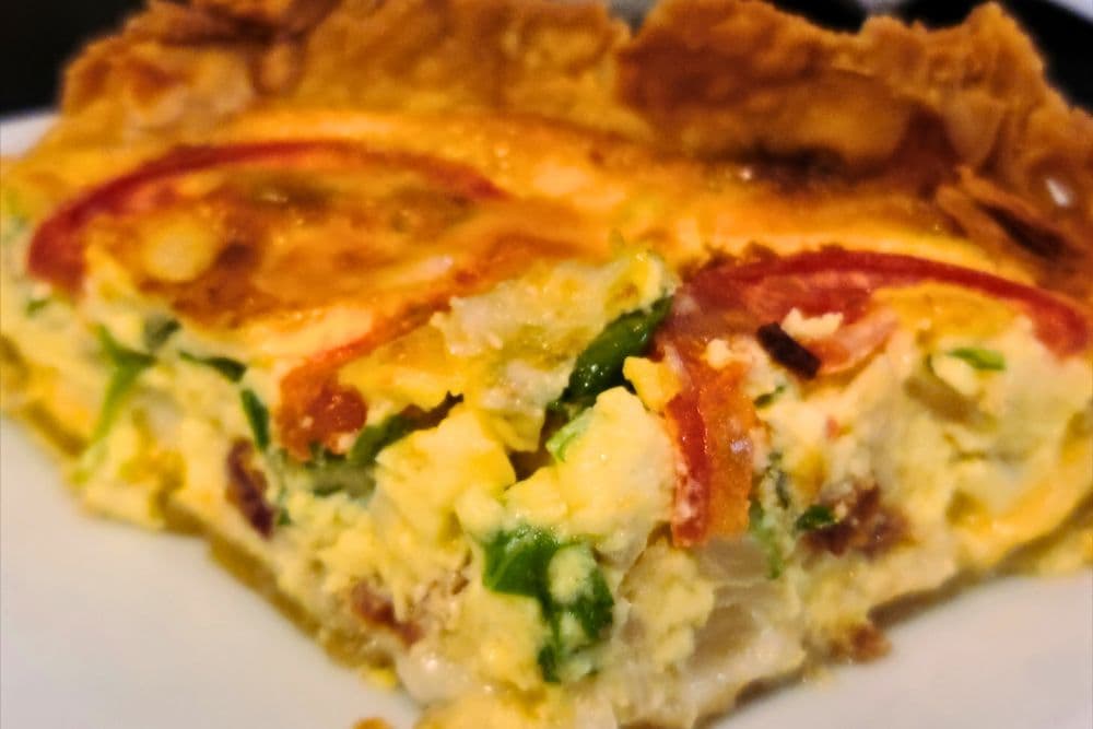 A slice of quiche with visible layers of egg, vegetables, and a flaky crust.
