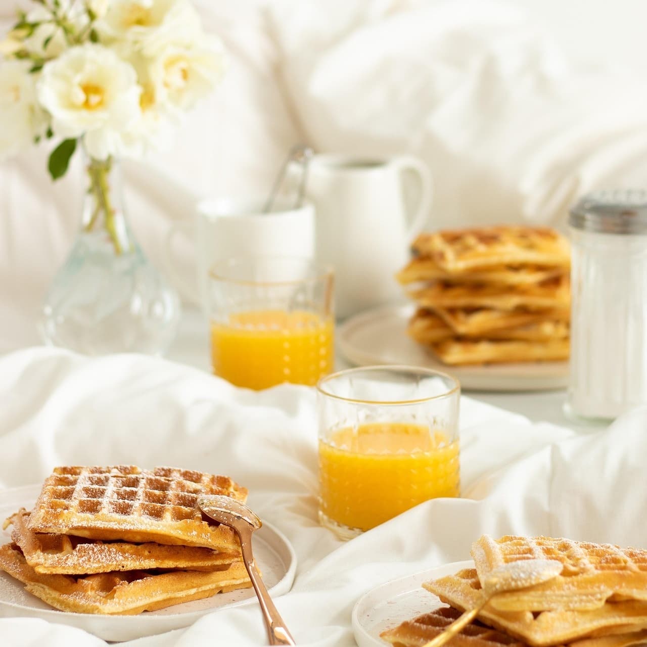 A cozy breakfast scene featuring waffles, orange juice, and fresh flowers on a bed.