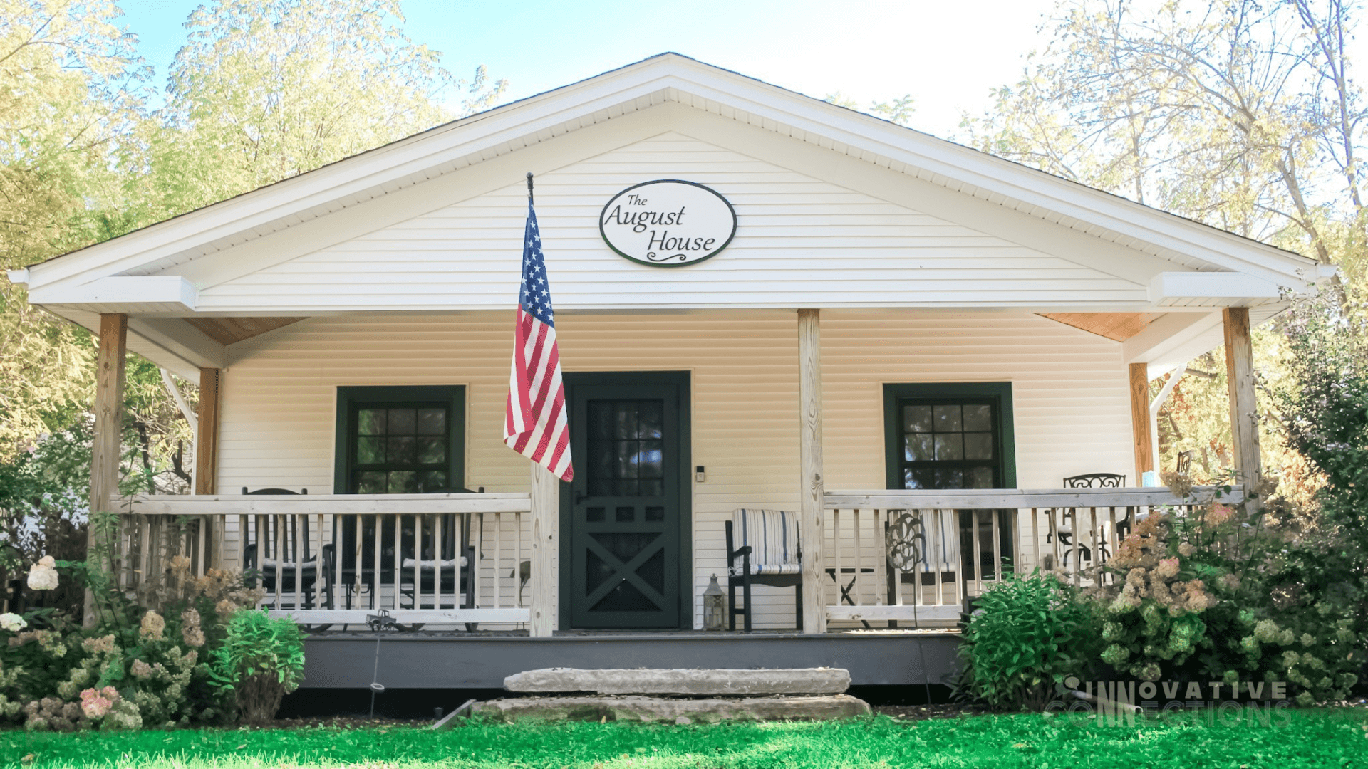 A charming white house with green accents, featuring a front porch, an American flag, and a sign reading "The August House."