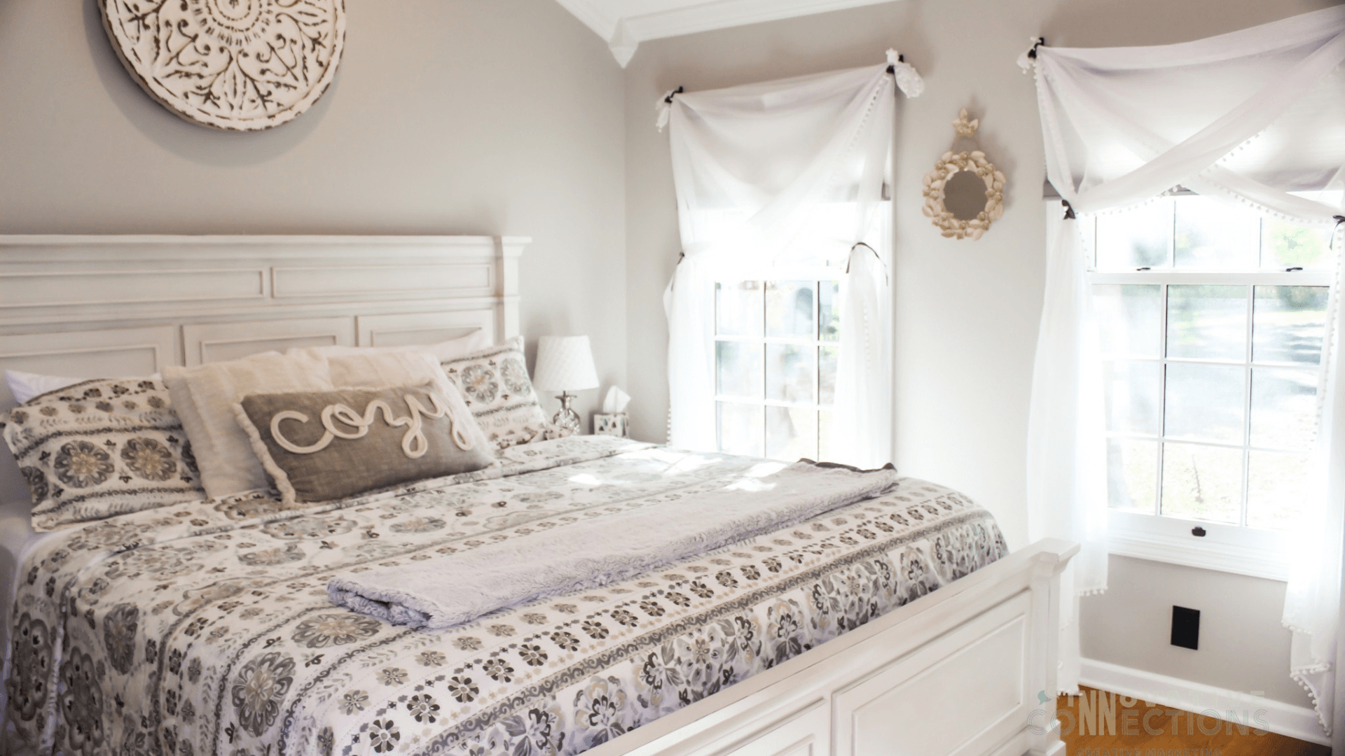 A bright and cozy bedroom with a white wooden bed, patterned bedding, and large windows adorned with sheer curtains.